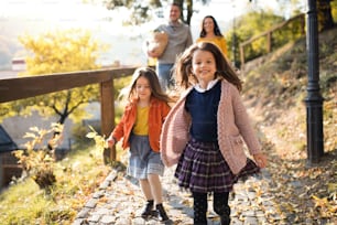 Deux filles avec des parents méconnaissables en arrière-plan marchant dans le parc en automne, se tenant la main.