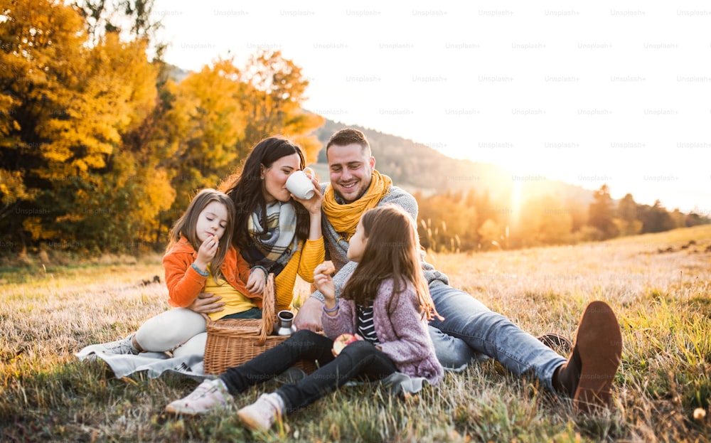 Ein Porträt einer glücklichen jungen Familie mit zwei kleinen Kindern, die bei Sonnenuntergang in der Herbstnatur auf einem Boden sitzen und ein Picknick machen.
