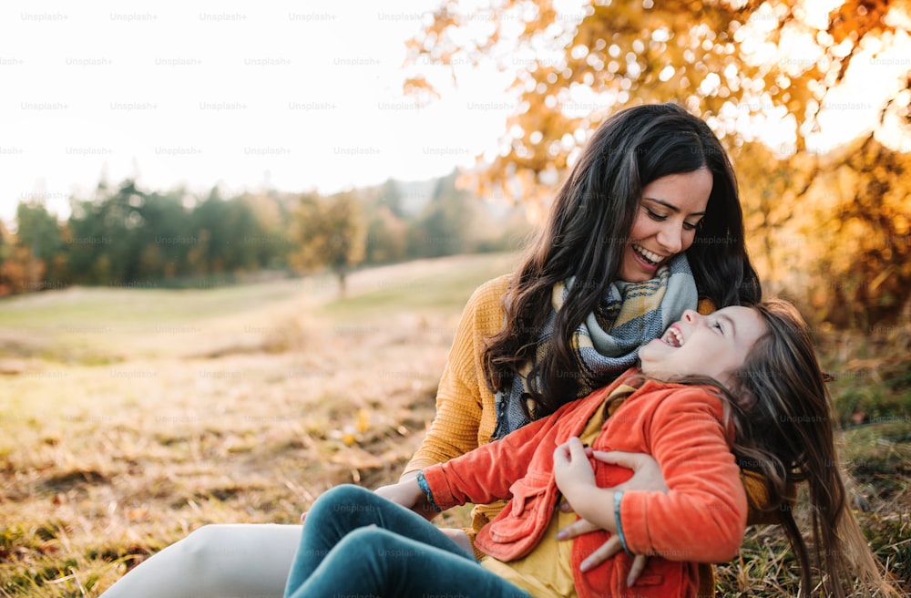 A portrait of young mother with a small daughter sitting on a ground in autumn nature at sunset.