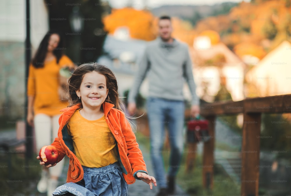 Kleines Mädchen mit nicht wiederzuerkennenden Eltern im Hintergrund, das im Herbst im Park läuft und Apfel isst.