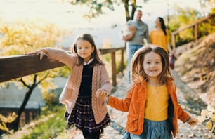 Duas meninas com pais irreconhecíveis ao fundo andando no parque no outono, de mãos dadas.
