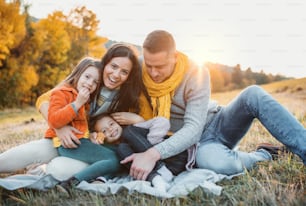 A portrait of happy young family with two small children sitting on a ground in autumn nature at sunset.