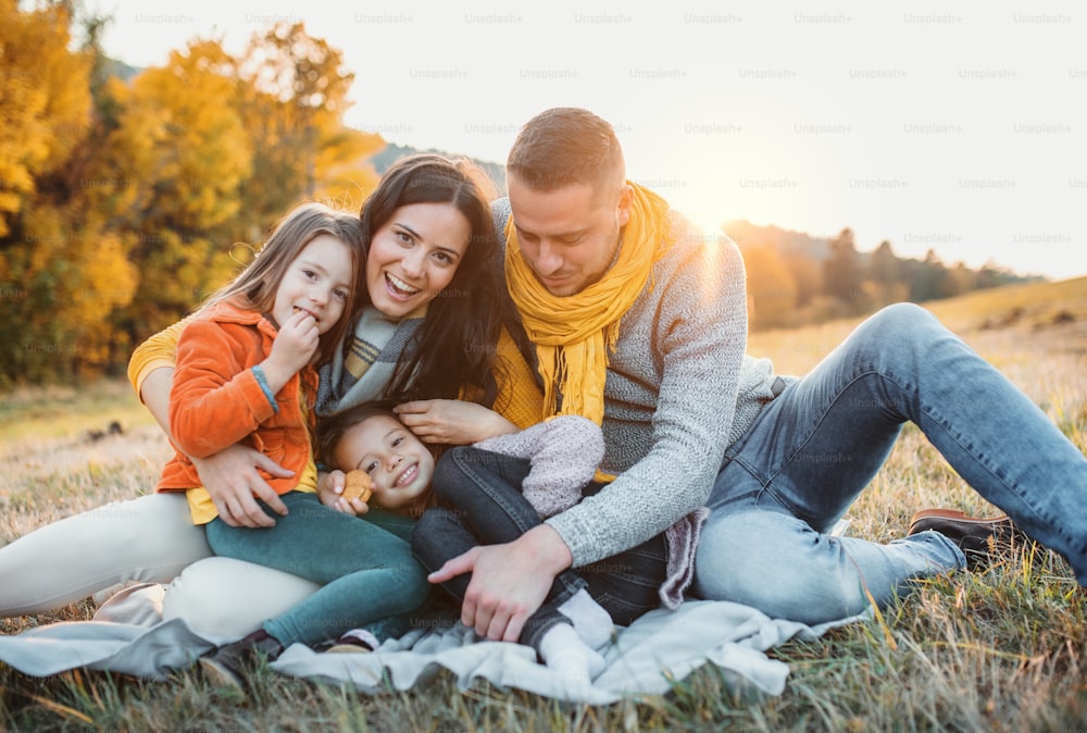 Ein Porträt einer glücklichen jungen Familie mit zwei kleinen Kindern, die bei Sonnenuntergang in der herbstlichen Natur auf einem Boden sitzen.