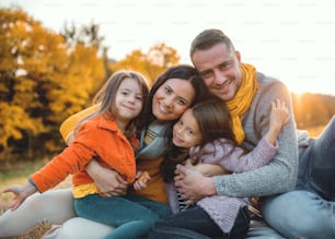 A portrait of happy young family with two small children sitting on a ground in autumn nature at sunset.