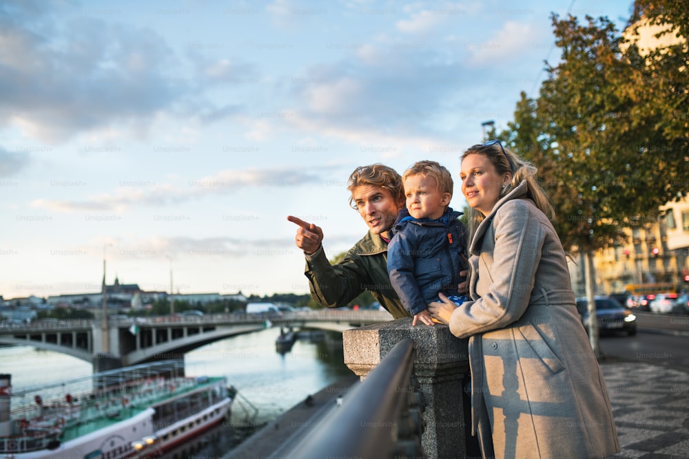 Padres jóvenes con su hijo pequeño parados al aire libre junto al río en la ciudad de Praga, señalando algo.