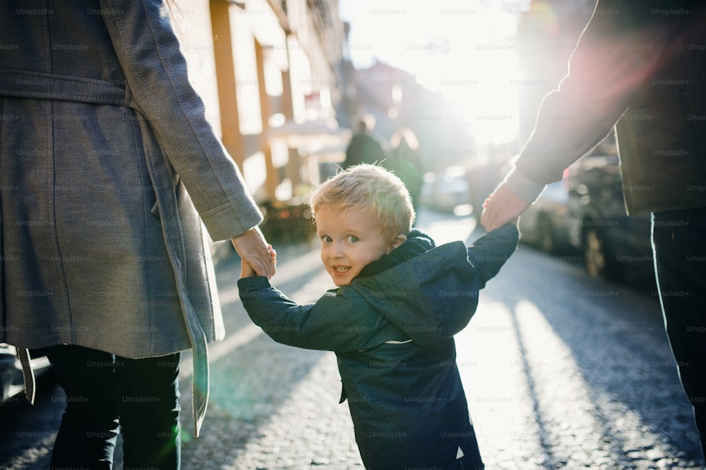 Una vista trasera de un niño pequeño con padres irreconocibles caminando al aire libre en la ciudad, tomados de la mano.