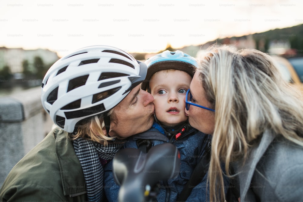 A small toddler boy with bicycle helmet and young parents outdoors in city, kissing.