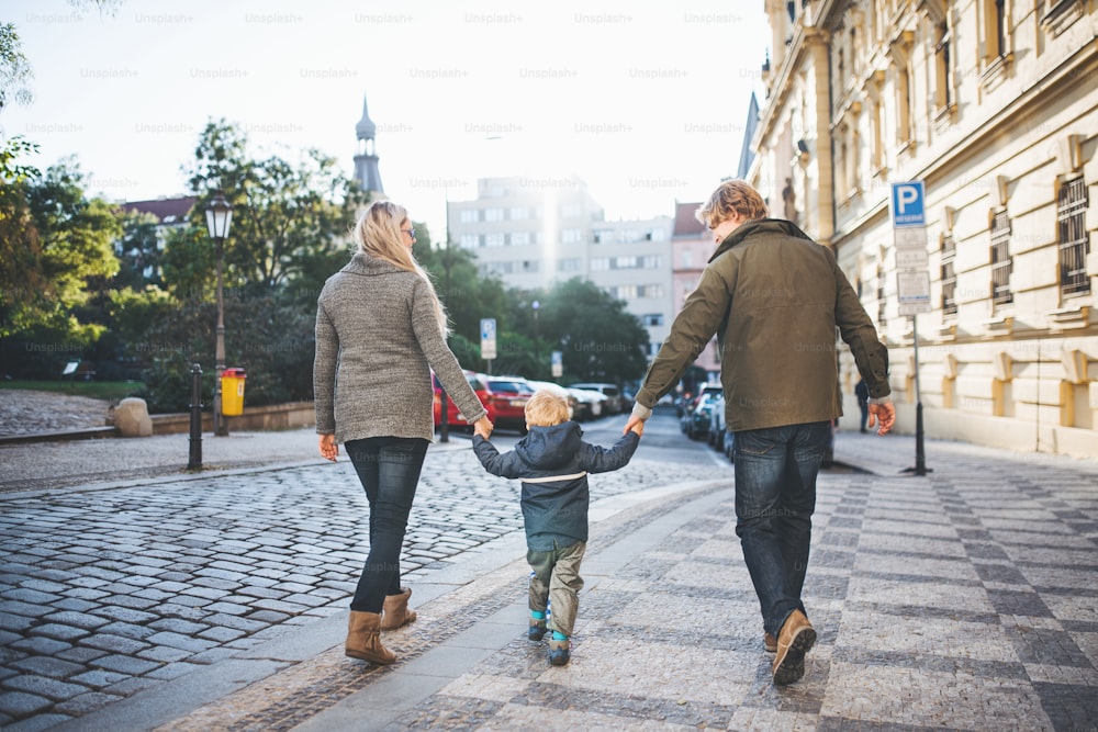Uma visão traseira de um menino pequeno com os pais andando ao ar livre na cidade, de mãos dadas.