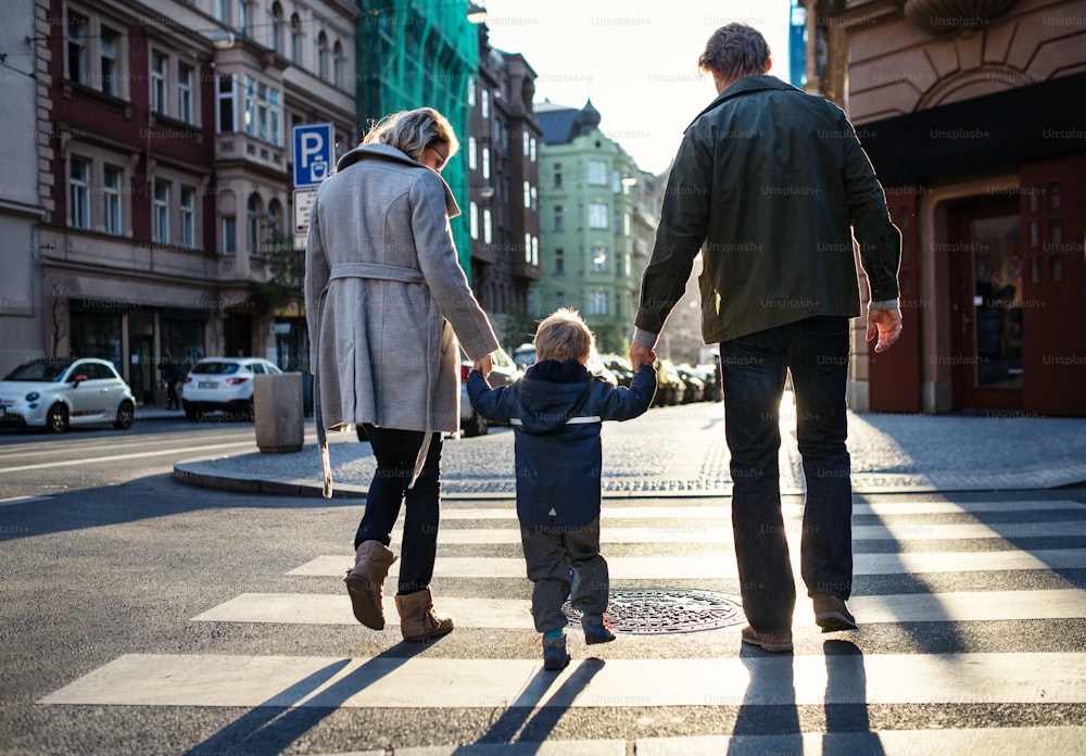 Uma visão traseira de um menino pequeno com os pais atravessando uma estrada ao ar livre na cidade, de mãos dadas.