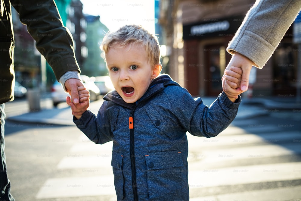 Um menino pequeno com pais irreconhecíveis andando ao ar livre na cidade, de mãos dadas.