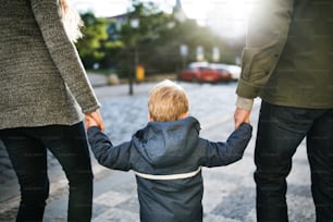A rear view of small toddler boy with unrecognizable parents walking outdoors in city, holding hands.