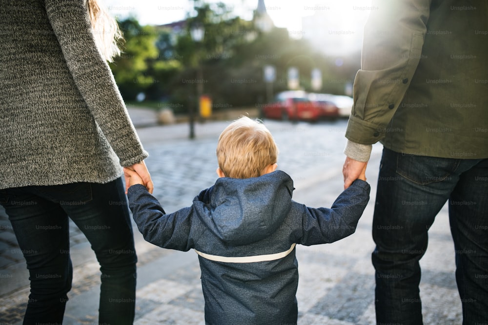 Uma visão traseira de um menino pequeno com pais irreconhecíveis andando ao ar livre na cidade, de mãos dadas.