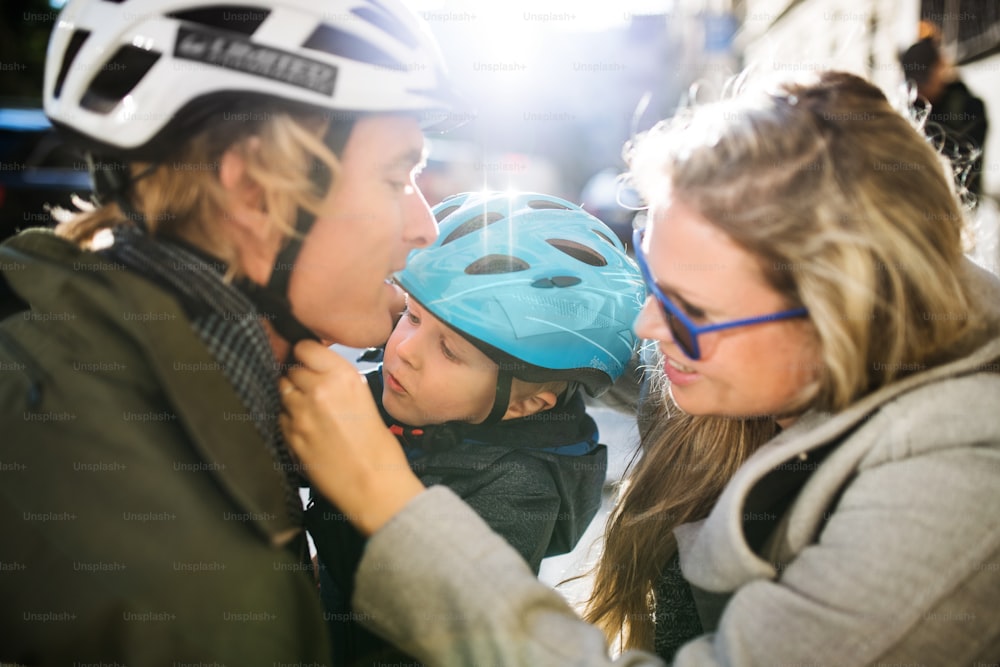 A small toddler boy with bicycle helmet and young parents outdoors in city.