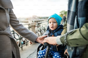 A small toddler boy with helmet sitting in bicycle seat with unrecognizable young parents outdoors on a street in city.