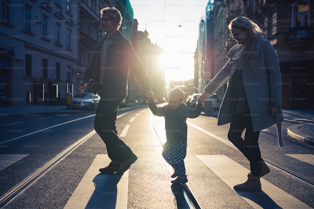 Un petit garçon en bas âge avec des parents traversant une route en plein air en ville au coucher du soleil, se tenant la main.