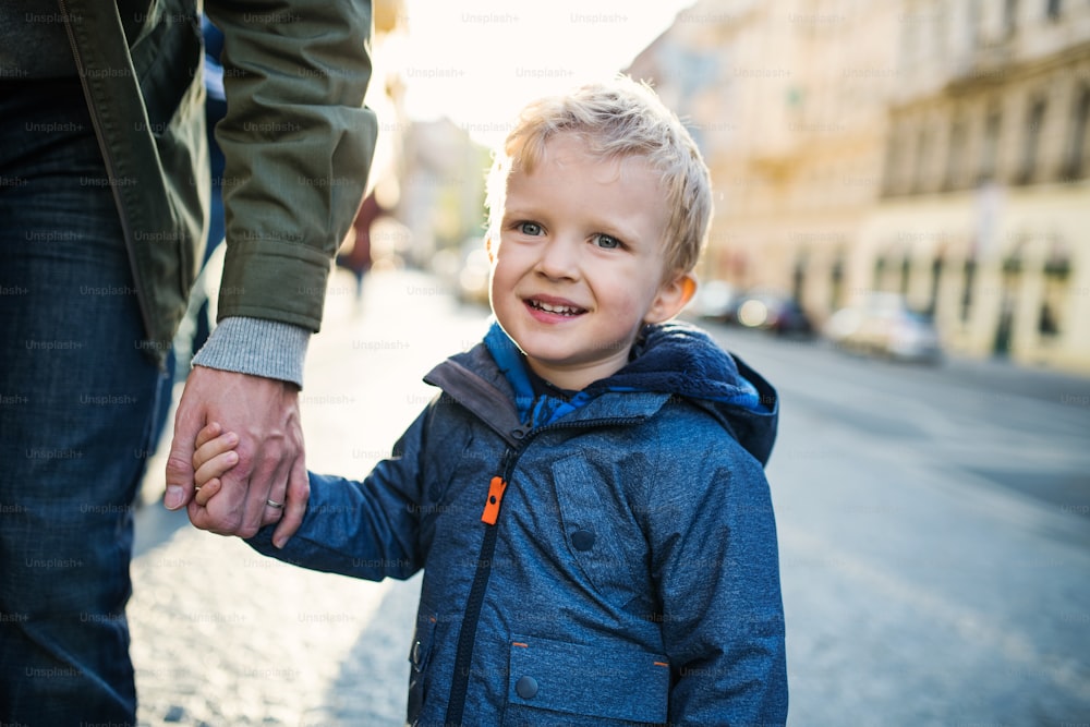 Um menino pequeno com pai irreconhecível andando ao ar livre na cidade, de mãos dadas.