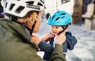 A young father putting on a bike helmet on his toddler son's head outdoors in city.