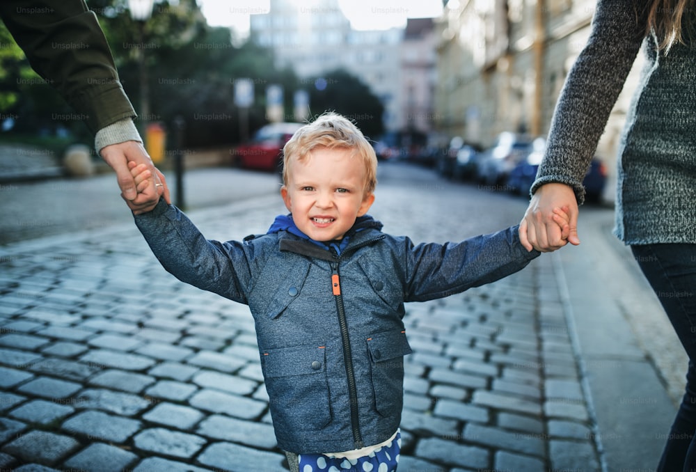 Un niño pequeño con padres irreconocibles caminando al aire libre en la ciudad, tomados de la mano.