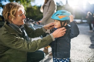 Un giovane padre che indossa un casco da bici sulla testa del figlio piccolo all'aperto in città.