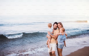 A happy young family with two toddler children standing on beach on summer holiday.