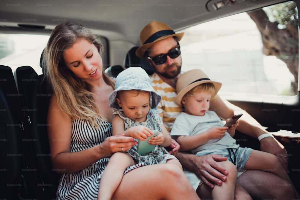 Une jeune famille avec deux enfants en bas âge dans une voiture de taxi pendant les vacances d’été, utilisant des smartphones.