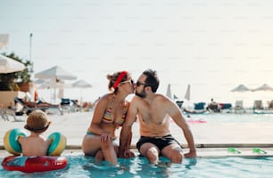 Un couple joyeux en maillot de bain assis au bord de la piscine pendant les vacances d’été, s’embrassant.