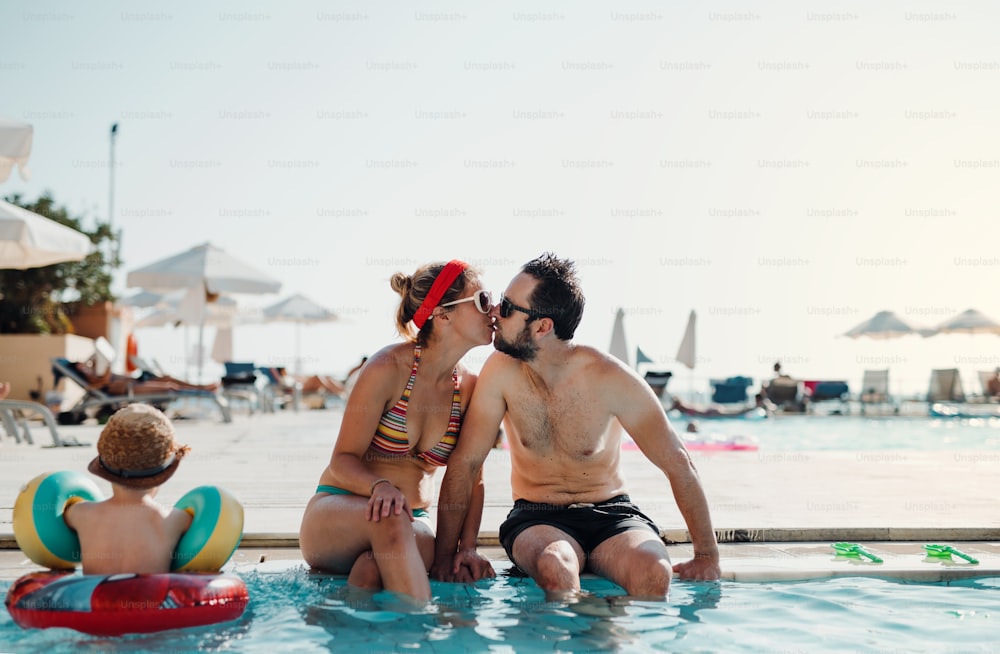 A cheerful couple in swimsuit sitting by the swimming pool on summer holiday, kissing.