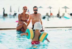 Ein kleines Kleinkind mit Armbinden und Eltern im Schwimmbad in den Sommerferien.