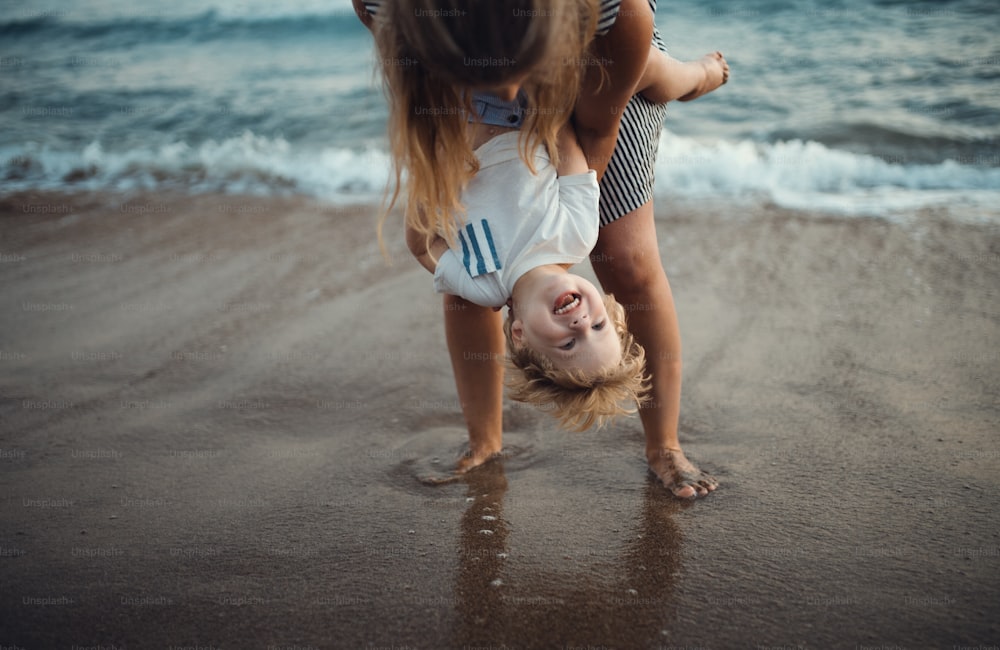 A young mother with a toddler boy standing on beach on summer holiday, having fun.
