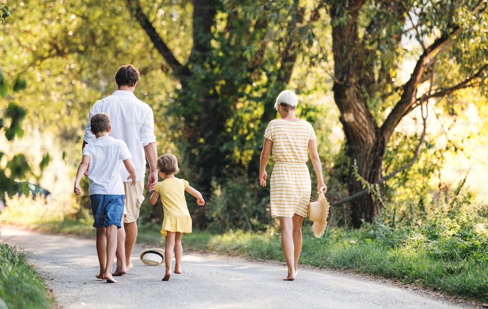 Uma visão traseira da família jovem com crianças pequenas andando descalças em uma estrada na natureza de verão.