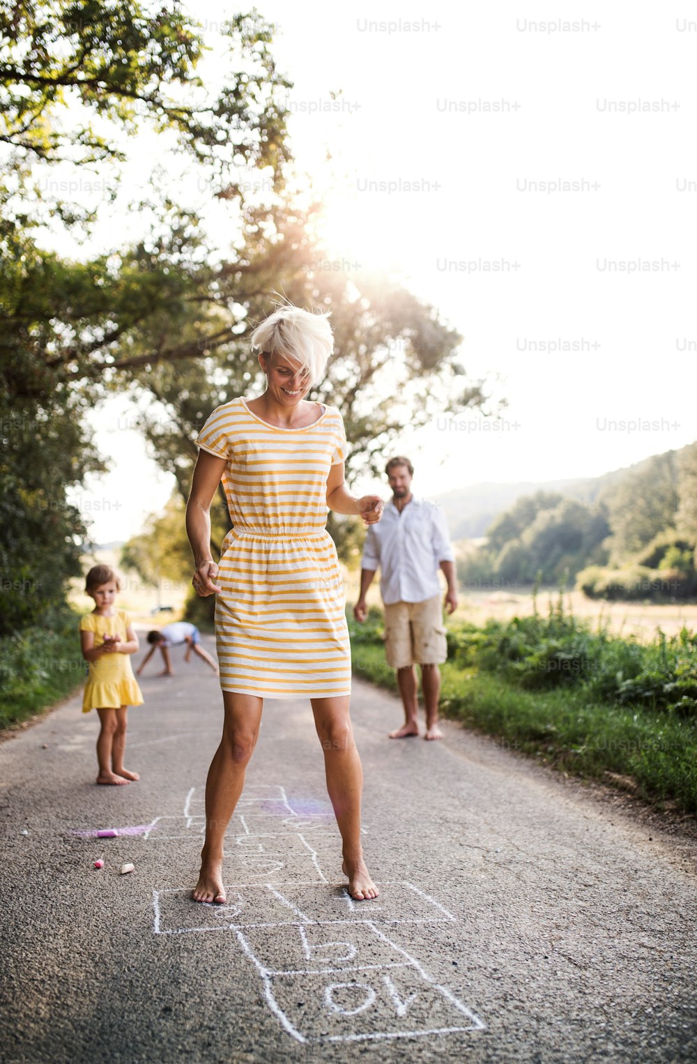 Una familia joven con niños pequeños jugando a la rayuela en una carretera en el campo en verano.
