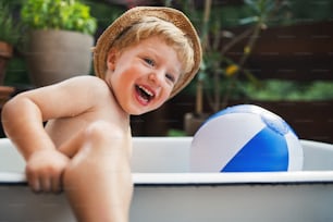 Happy small boy with a hat in bath tub outdoors in garden in summer, playing in water.