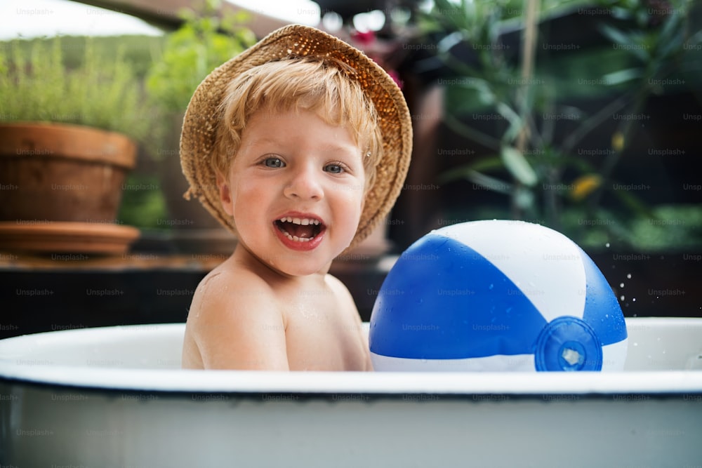 Happy small boy with a ball in bath tub outdoors in garden in summer, playing in water.