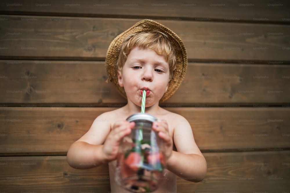 A front view of small boy with a drink standing against dark wooden background on a patio in summer. Copy space.