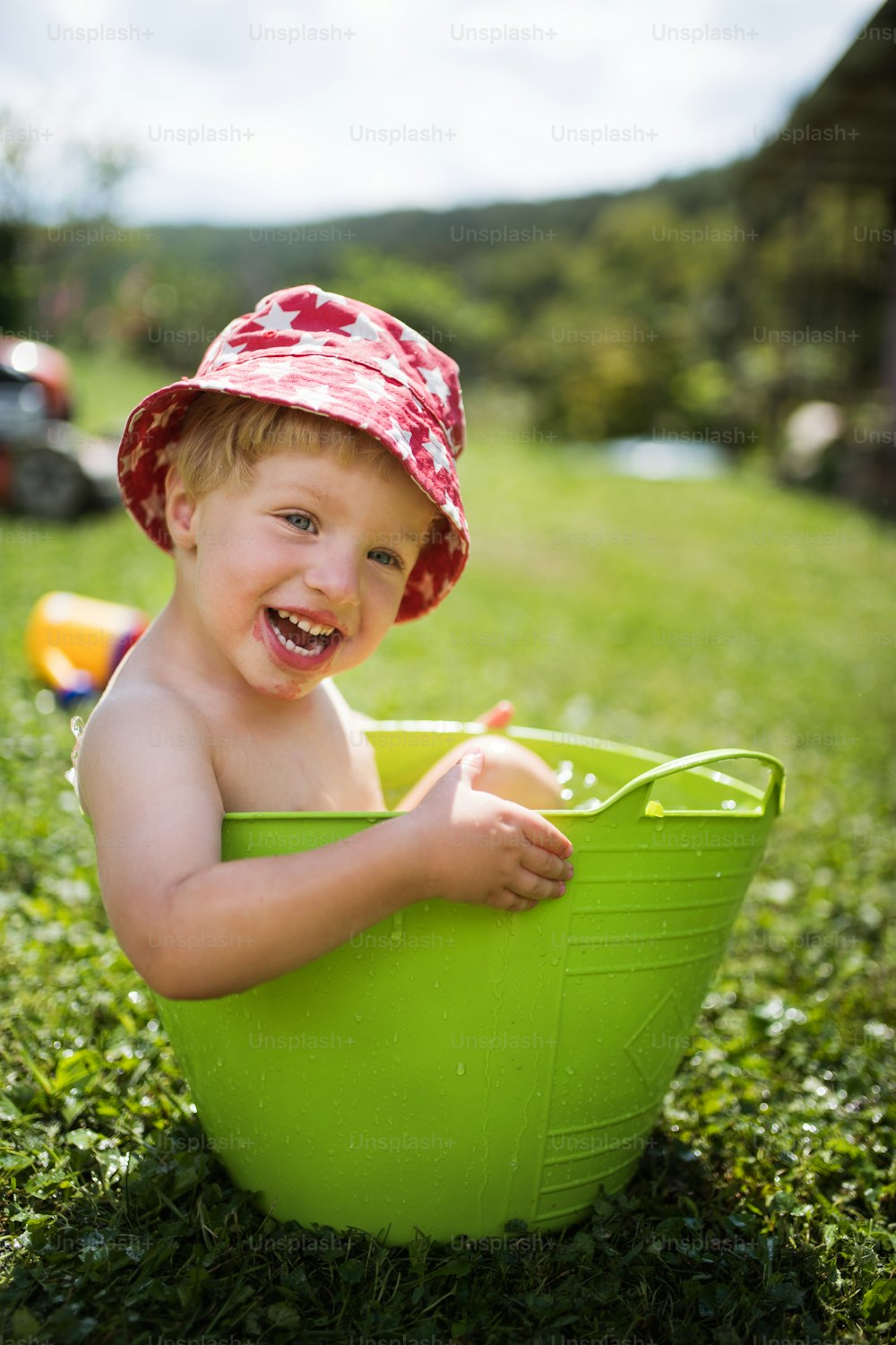 Small boy with a hat sitting in plastic bucket outdoors in garden in summer, playing in water.