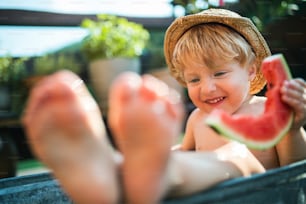 Happy small boy with a hat in bath tub outdoors in garden in summer, eating watermelon.