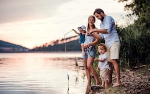 Eine junge Familie mit zwei Kleinkindern im Sommer draußen am Fluss und spielt mit Papierbooten.