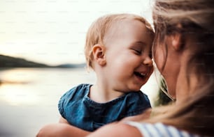 A close-up of mother with a toddler daughter outdoors by the river in summer, laughing.