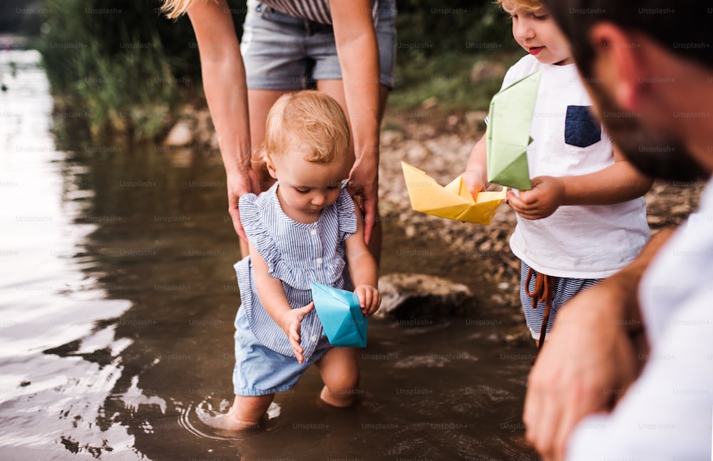 Una sección media de una familia joven con dos niños pequeños al aire libre junto al río en verano, jugando con botes de papel.