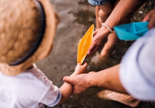 A midsection of young family with toddler child outdoors by the river in summer, playing with paper boats.