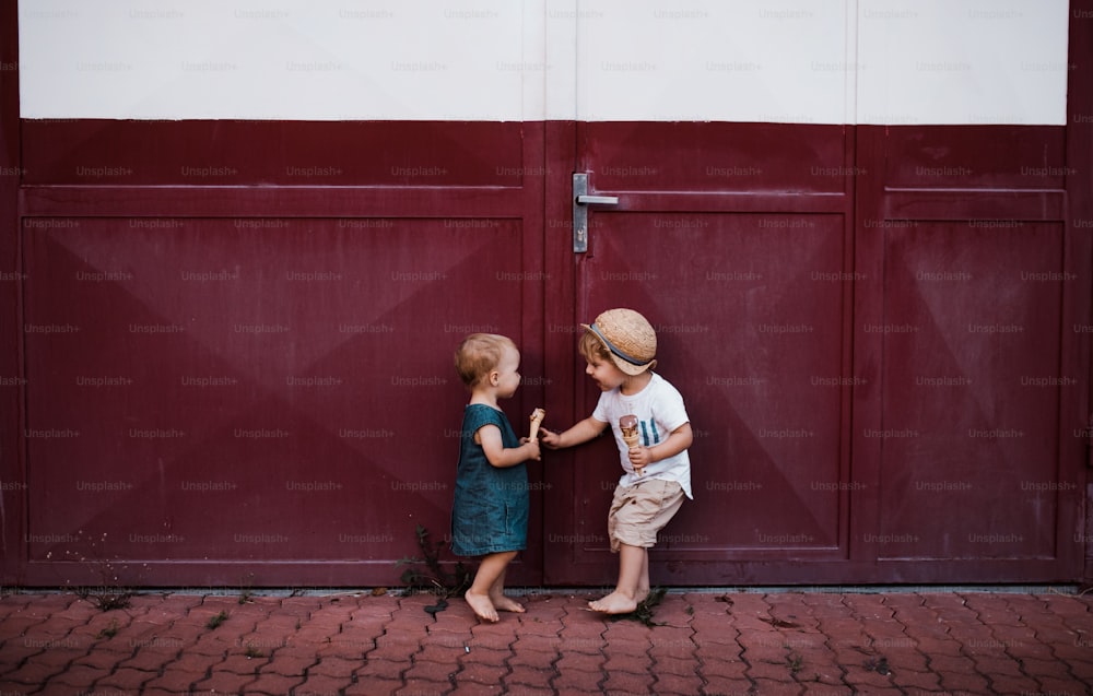 Two small toddler children outdoors in summer, eating ice cream and standing in front of a gate.