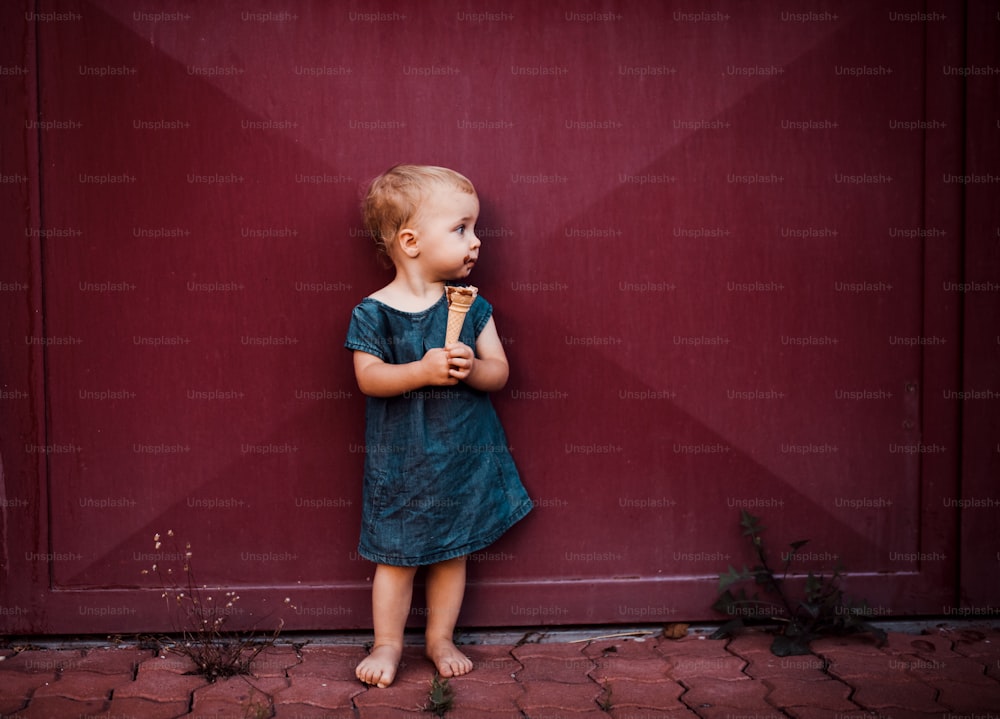 A front view of cute small toddler girl outdoors in summer, eating ice cream.