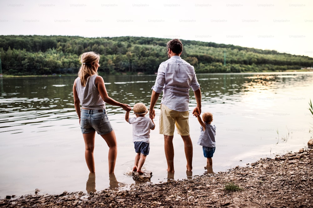Una vista trasera de una familia joven con dos niños pequeños que pasan tiempo al aire libre junto al río en verano.