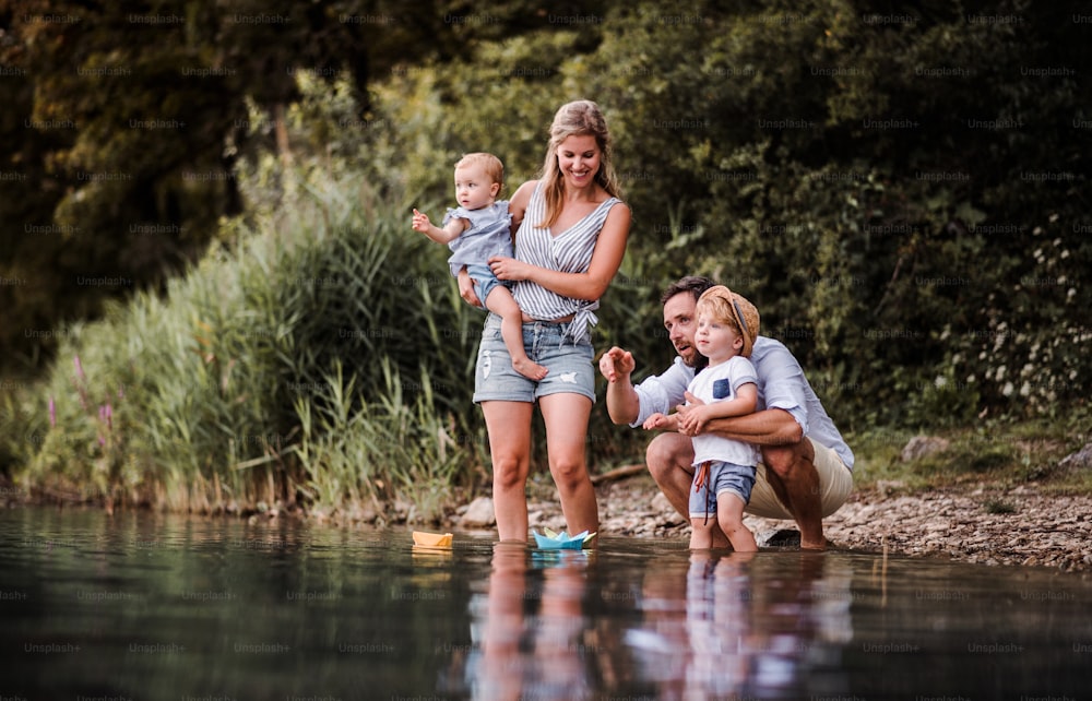 Eine junge Familie mit zwei Kleinkindern im Sommer draußen am Fluss und spielt mit Papierbooten.