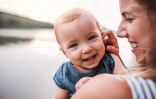 A close-up of mother with a toddler daughter outdoors by the river in summer, laughing.