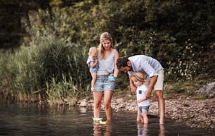 A young family with two toddler children outdoors by the river in summer, playing with paper boats.