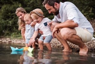 A young family with two toddler children outdoors by the river in summer, playing with paper boats.