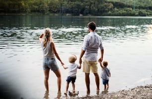 A rear view of young family with two toddler children spending time outdoors by the river in summer.