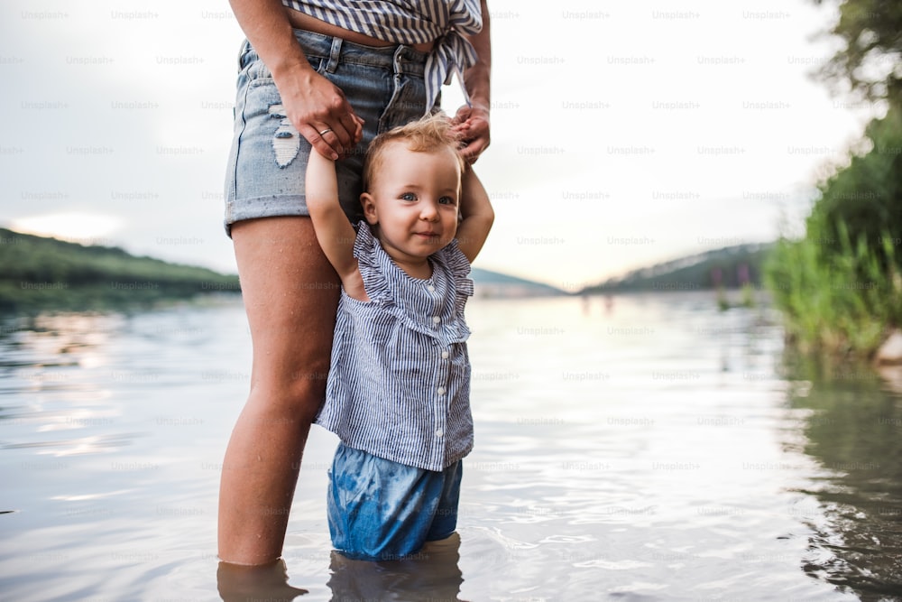 Uma barriga de mãe com uma filha pequena ao ar livre à beira do rio no verão, em pé na água.