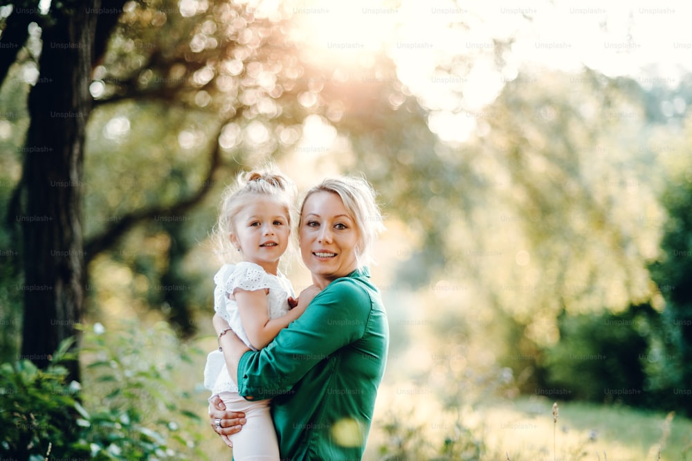 Beautiful young mother in green sunny summer nature holding her cute small daughter in the arms. Copy space.